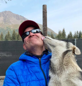man wearing sunglasses and a blue coat being licked by a husky